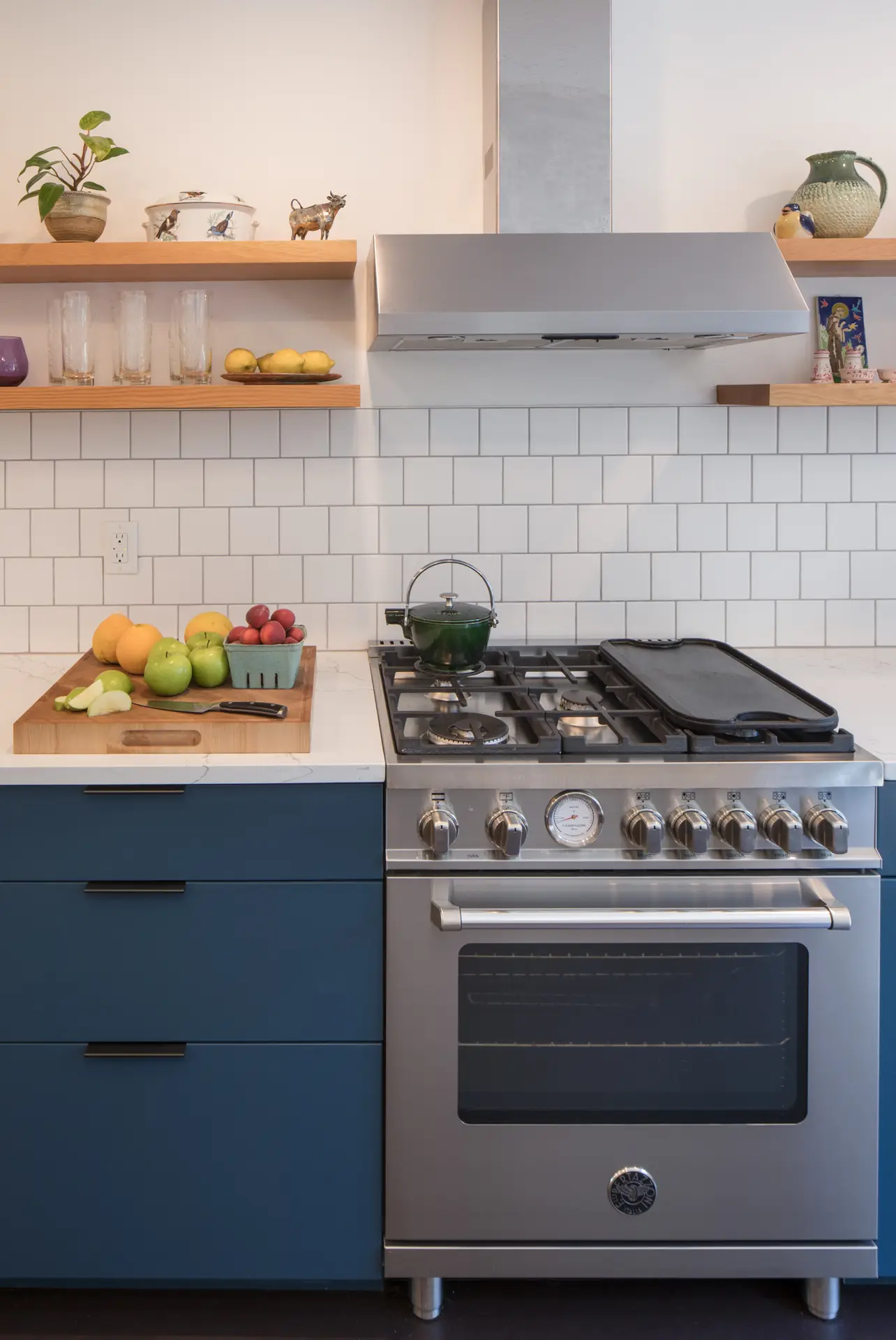 Little Larch kitchen, featuring the range and range hood, flanked by warm wood floating shelves and navy blue lower cabinets.