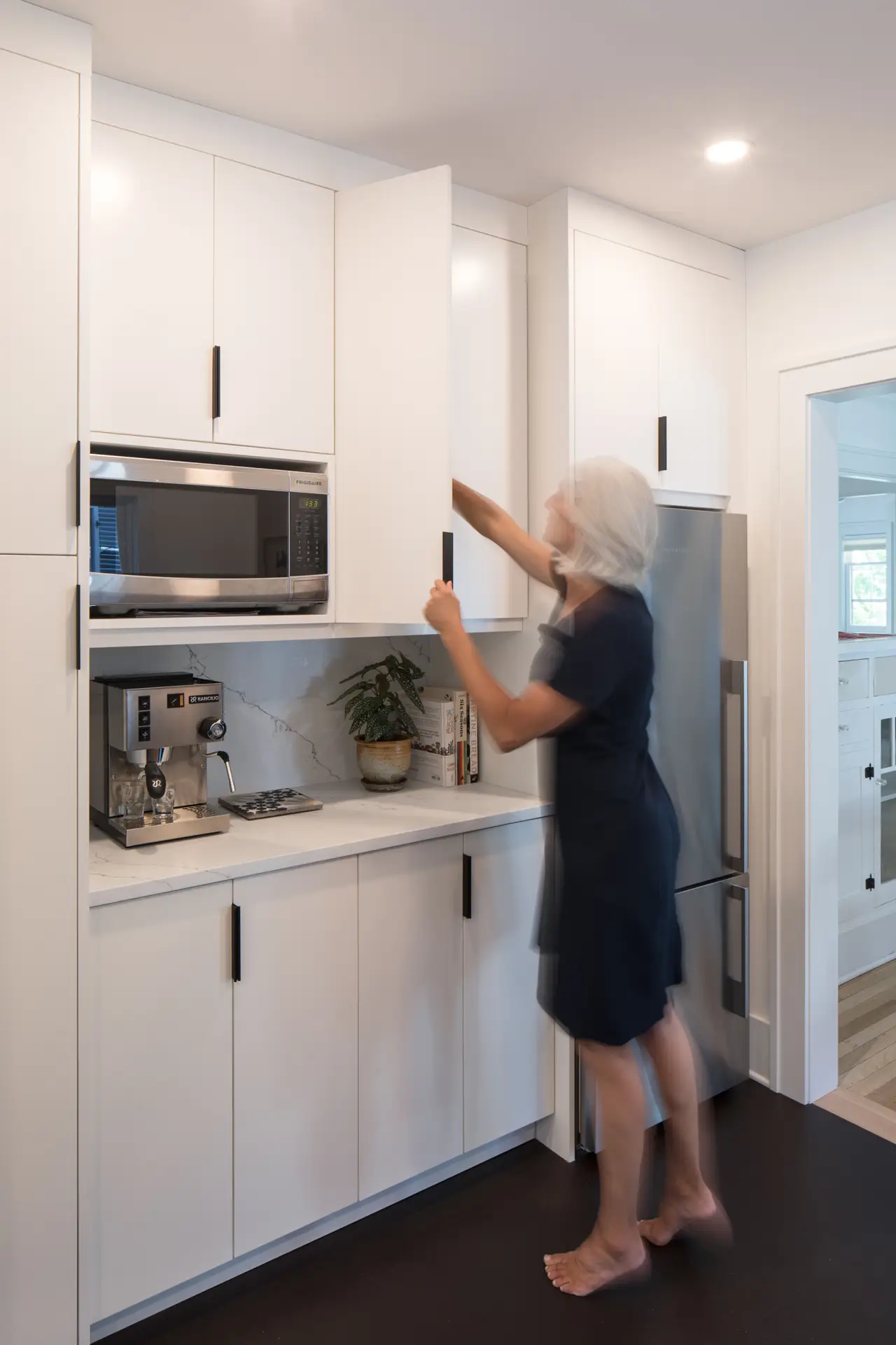 Little Larch kitchen, featuring white upper and lower cabinets and a woman pulling something out of the upper cabinets.