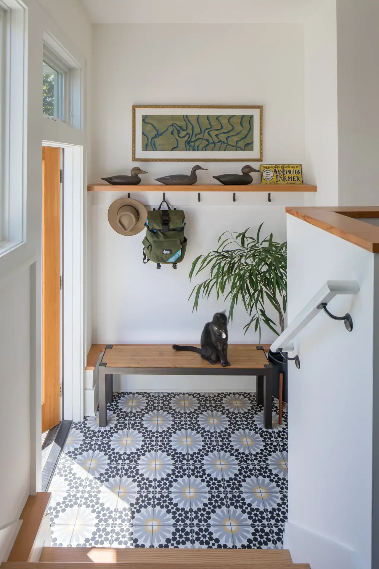 Little Larch entry, featuring a playful and visually exciting tile floor. The floor is various shades of blue and has a floral geometric pattern. The space also features a bench for removing shoes and upper shelf with coat hooks. Several decorative wood ducks are perched atop the shelf.