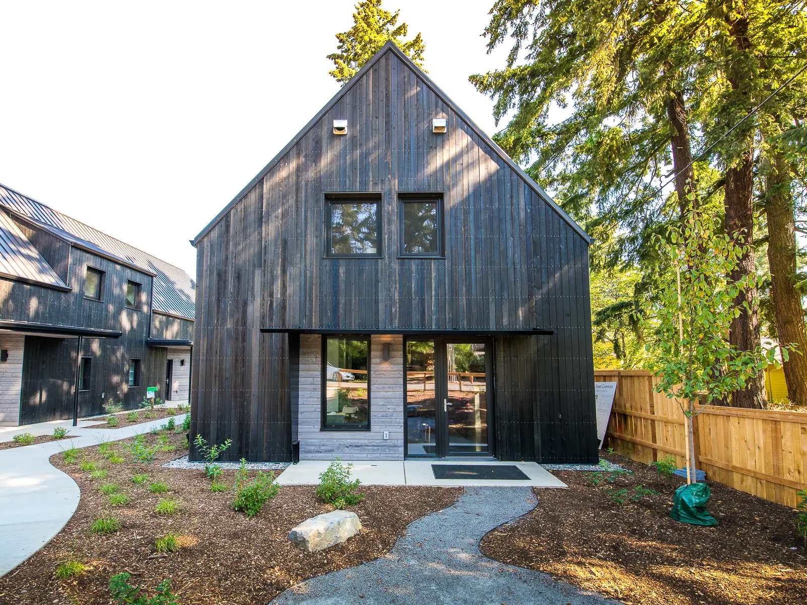 The southern exposure of a duplex at Going Street Commons. The structure features a prominent, steep-gabled roof and a minimalist silhouette, which is a modern interpretation of traditional barn-style architecture. The exterior is clad in Pioneer Millworks Larch Siding with a "Black" finish, achieved through a charring process similar to the Japanese shou sugi ban technique. This finish is both durable and rot-resistant.