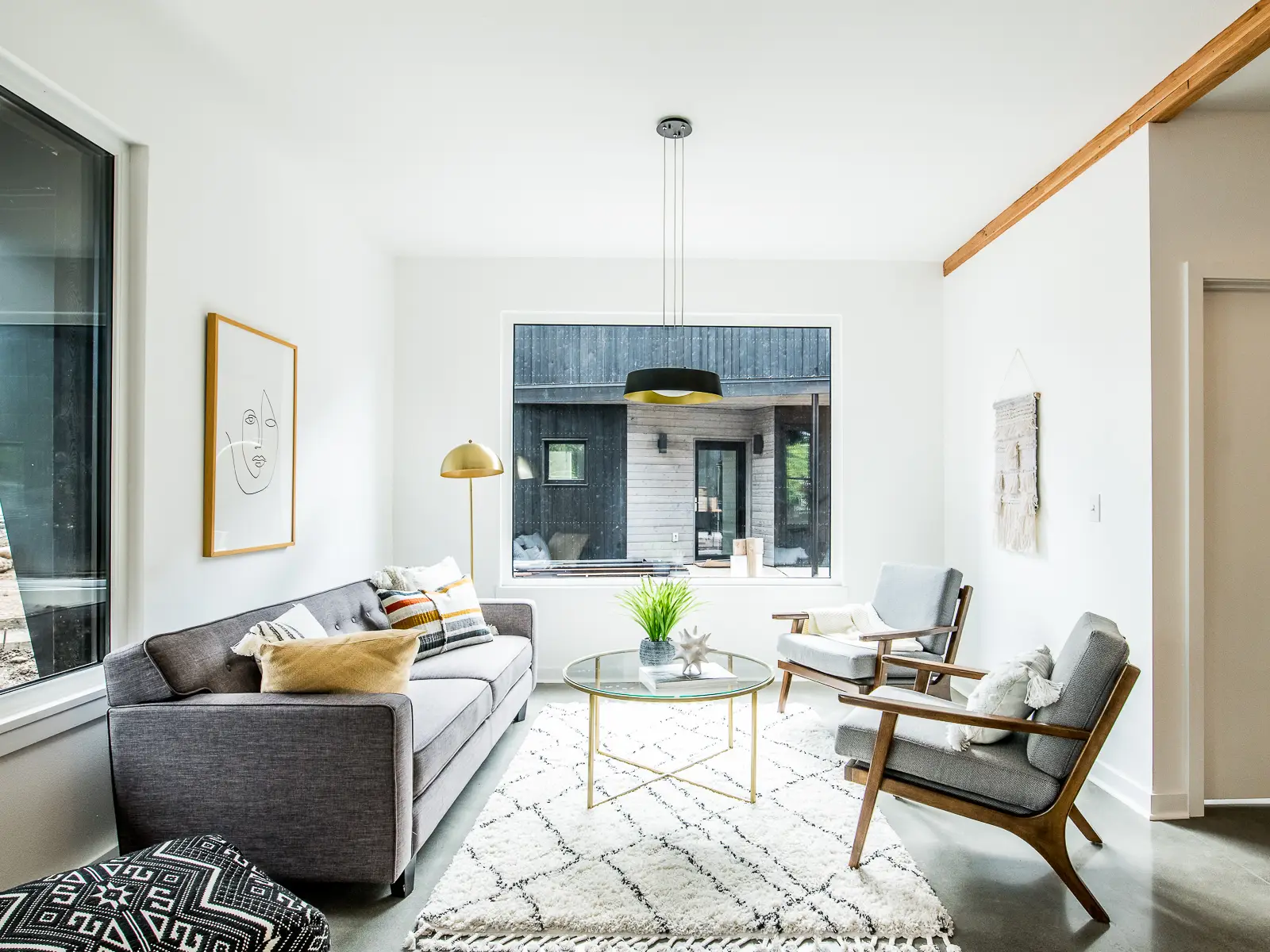 A bright, mid-century modern living room of a home in Going Street Commons. The space features a grey tufted sofa paired with two Z-style wooden armchairs upholstered in grey fabric. A black-and-white geometric patterned pouf sits in the foreground, adding an extra layer of texture. The room is characterized by polished concrete floors and crisp white walls. A large, natural wood ceiling beam provides a warm, structural accent.