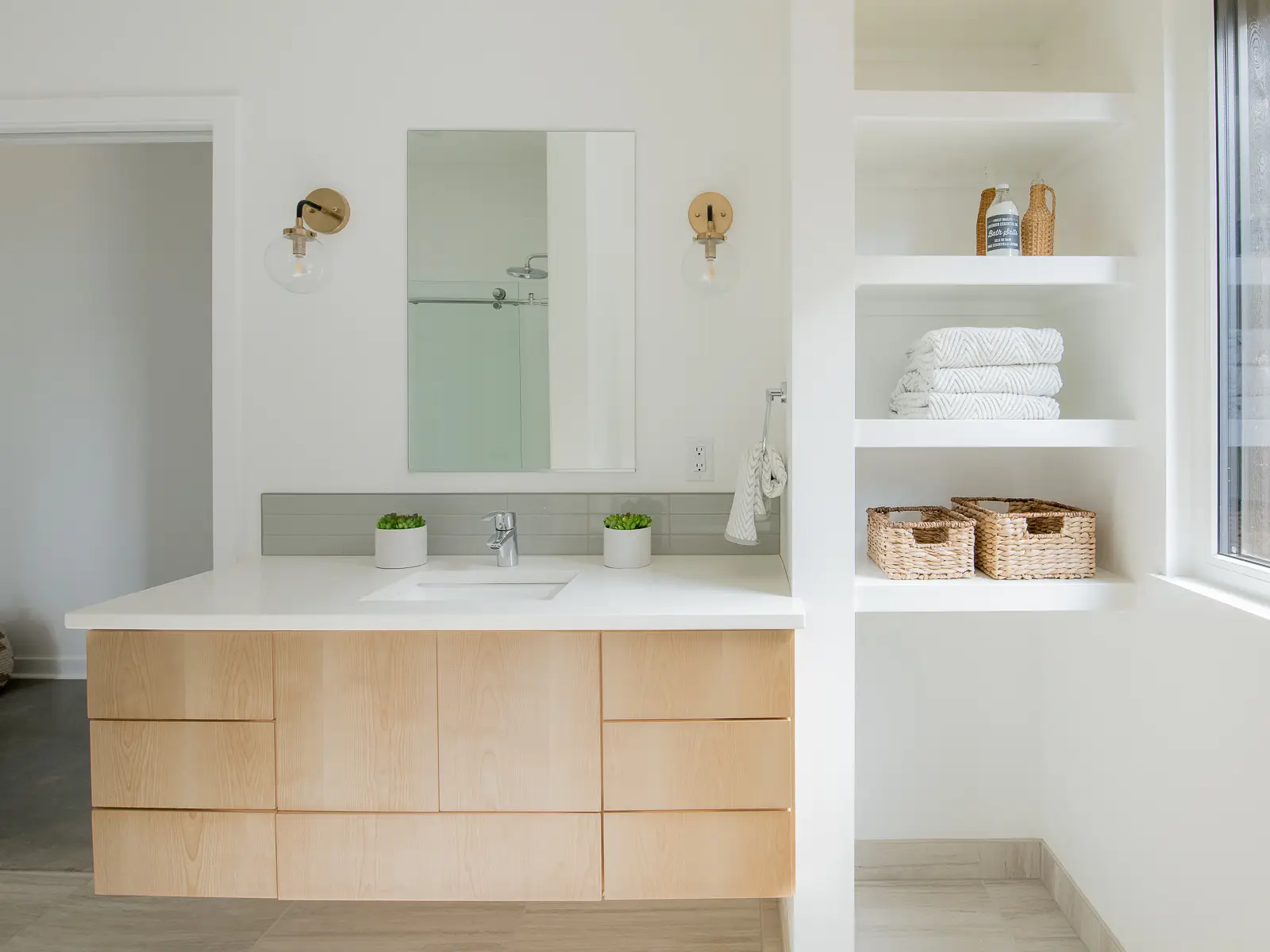A floating light wood vanity with a clean, handle-less design. The floating installation creates an airy feel and makes the floor easier to clean. To the right, a floor-to-ceiling recessed shelving unit provides ample open storage for towels and decorative baskets, finished in a crisp white that blends into the wall.