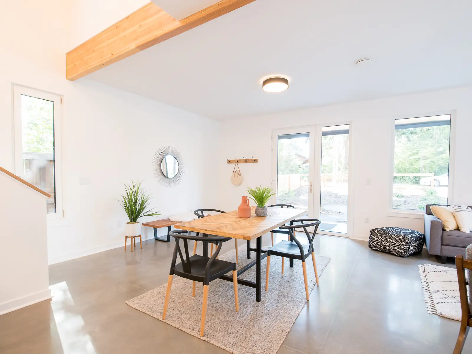 A bright, open-concept interior of a home at Going Street Commons. The focal point is a custom chevron-patterned wood dining table paired with six black wishbone-style chairs. The arrangement sits atop a light grey textured area rug, creating a defined space within the open floor plan. The room features sleek, polished concrete floors, which provide a durable and industrial contrast to the crisp white walls and warm wood accents. A large, exposed natural wood beam spans the ceiling, adding structural character.