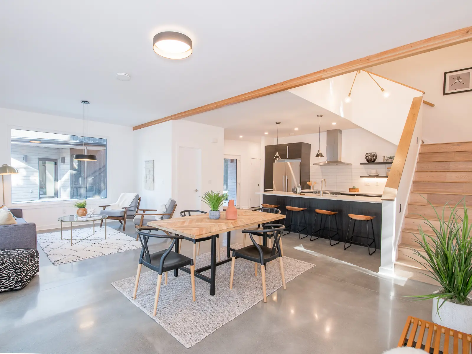 An open-plan first floor of a home at Going Street Commons. The background features a sleek kitchen with a black island, wood-topped bar stools, and white subway tile backsplash. It includes professional-grade stainless steel appliances and a chimney-style vent hood.