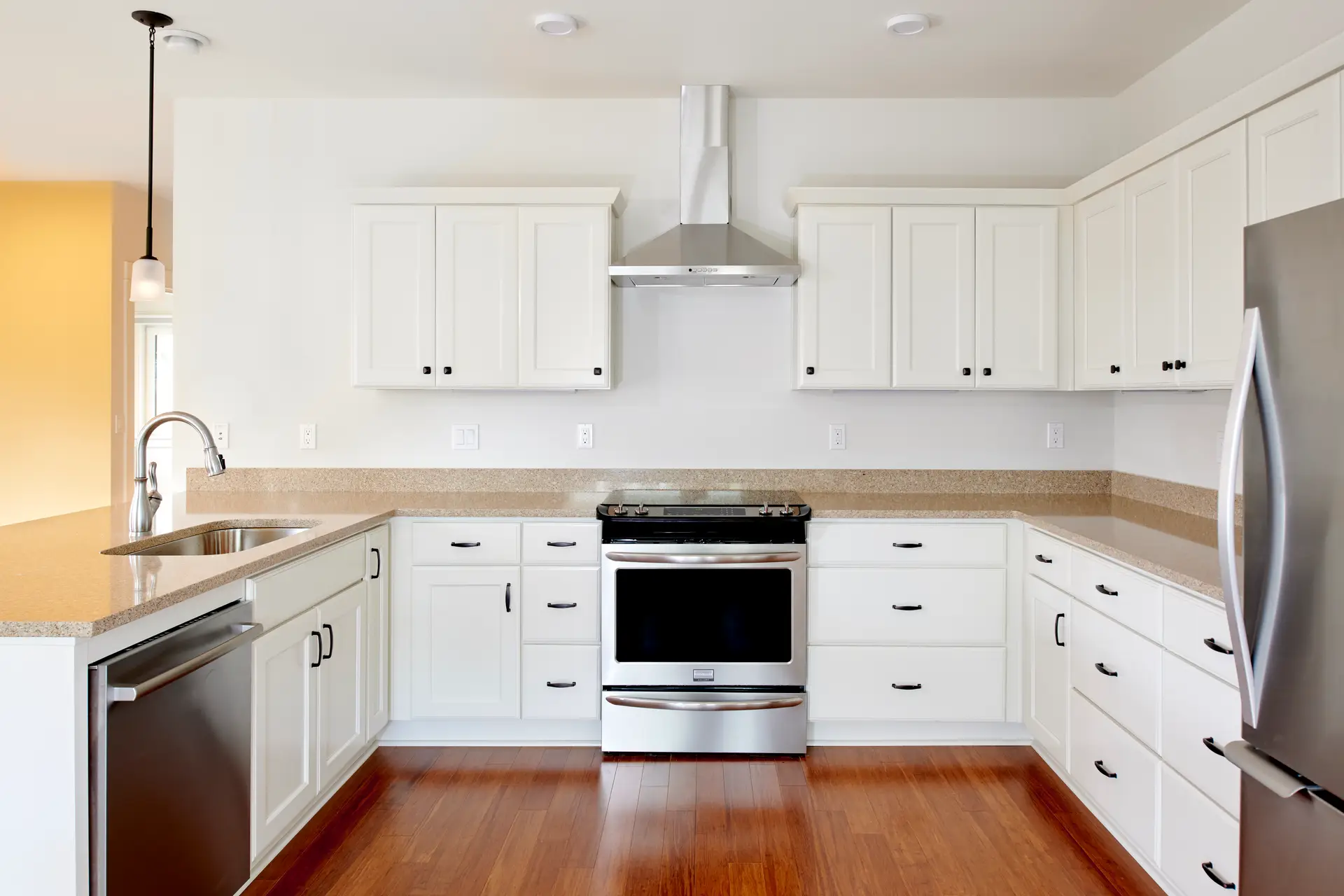 The Oaks at Rose Villa kitchen, highlighting the light and bright walls and cabinets that are centered around a range and range hood in the center of the frame. A fridge frames the right side and a yellow wall in the distance frames the left side along with a sink and dishwasher in the peninsula in the left of frame.