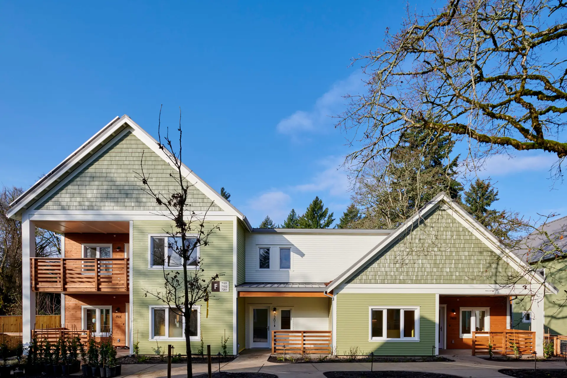 The Oaks at Rose Villa from the exterior. The building reflects the craftsman style of the neighborhood and features light green horizontal cladding combined with slightly darker green shake siding at the gable ends. There are also set back porches that feature warm wood cladding.