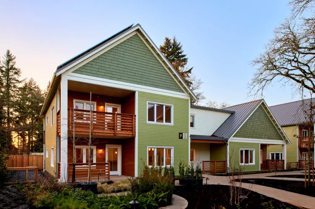 The Oaks at Rose Villa from the exterior. The building reflects the craftsman style of the neighborhood and features light green horizontal cladding combined with slightly darker green shake siding at the gable end. There are also set back porches that feature warm wood cladding.