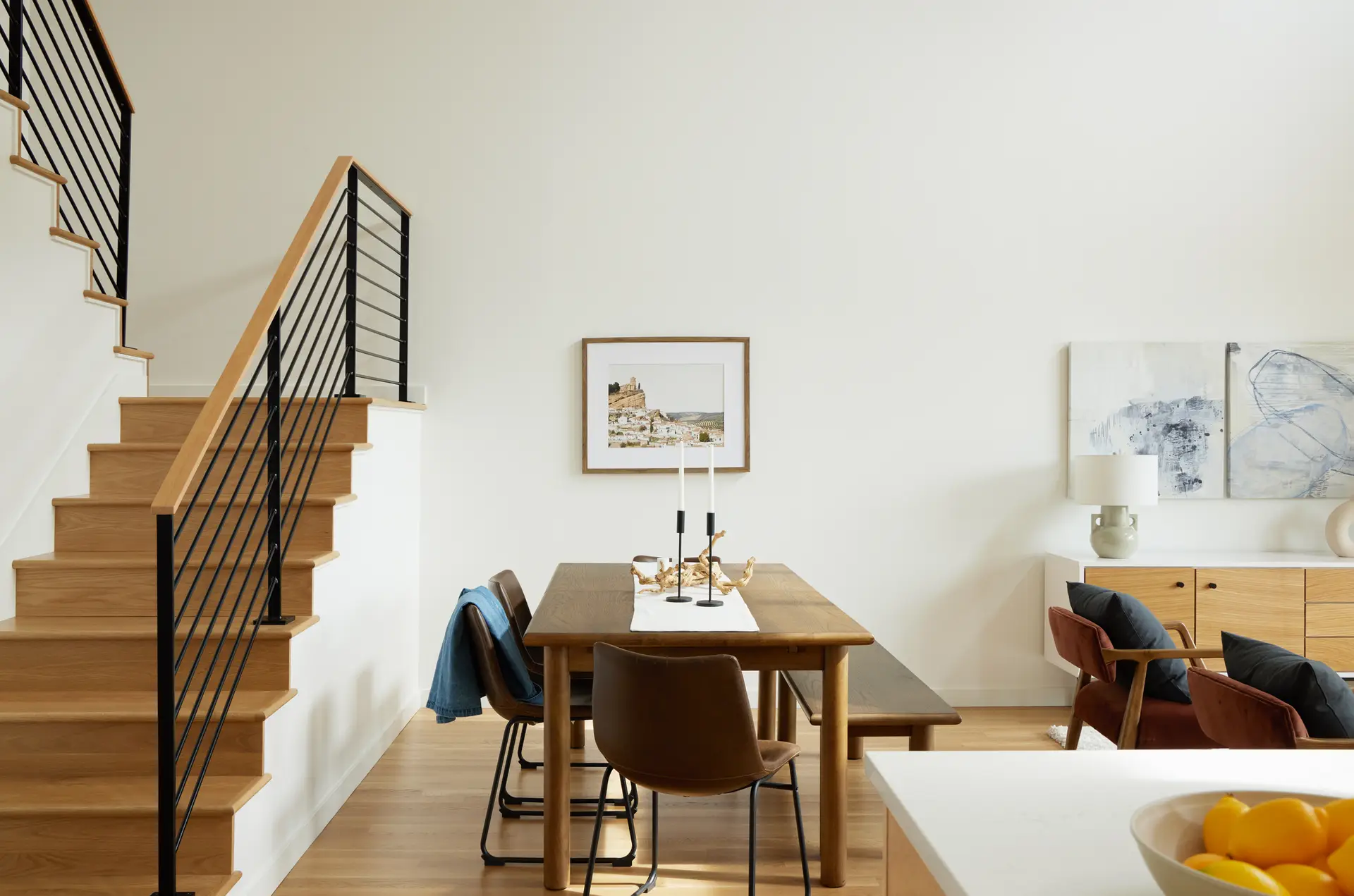 An open-concept living and dining area of an "urban loft" style townhome at Trillium Townhomes, a zero-energy community. A simple, mid-century modern wooden dining table is paired with brown leather chairs and a matching wooden bench. It features a minimalist centerpiece of two thin black candlesticks on a white runner. A prominent wooden staircase with a black metal cable railing leads to the second floor. The high, white walls and light-wood flooring contribute to a bright and airy atmosphere.