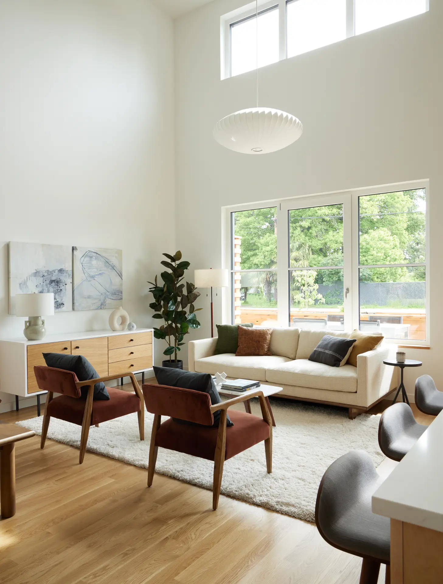 Light-filled, double-height living room of a residence at Trillium Townhomes. The room is characterized by its soaring double-height ceilings and crisp white walls, which amplify the sense of openness and light. A large, saucer-shaped Nelson Bubble Lamp hangs prominently from the high ceiling, serving as a mid-century modern focal point.