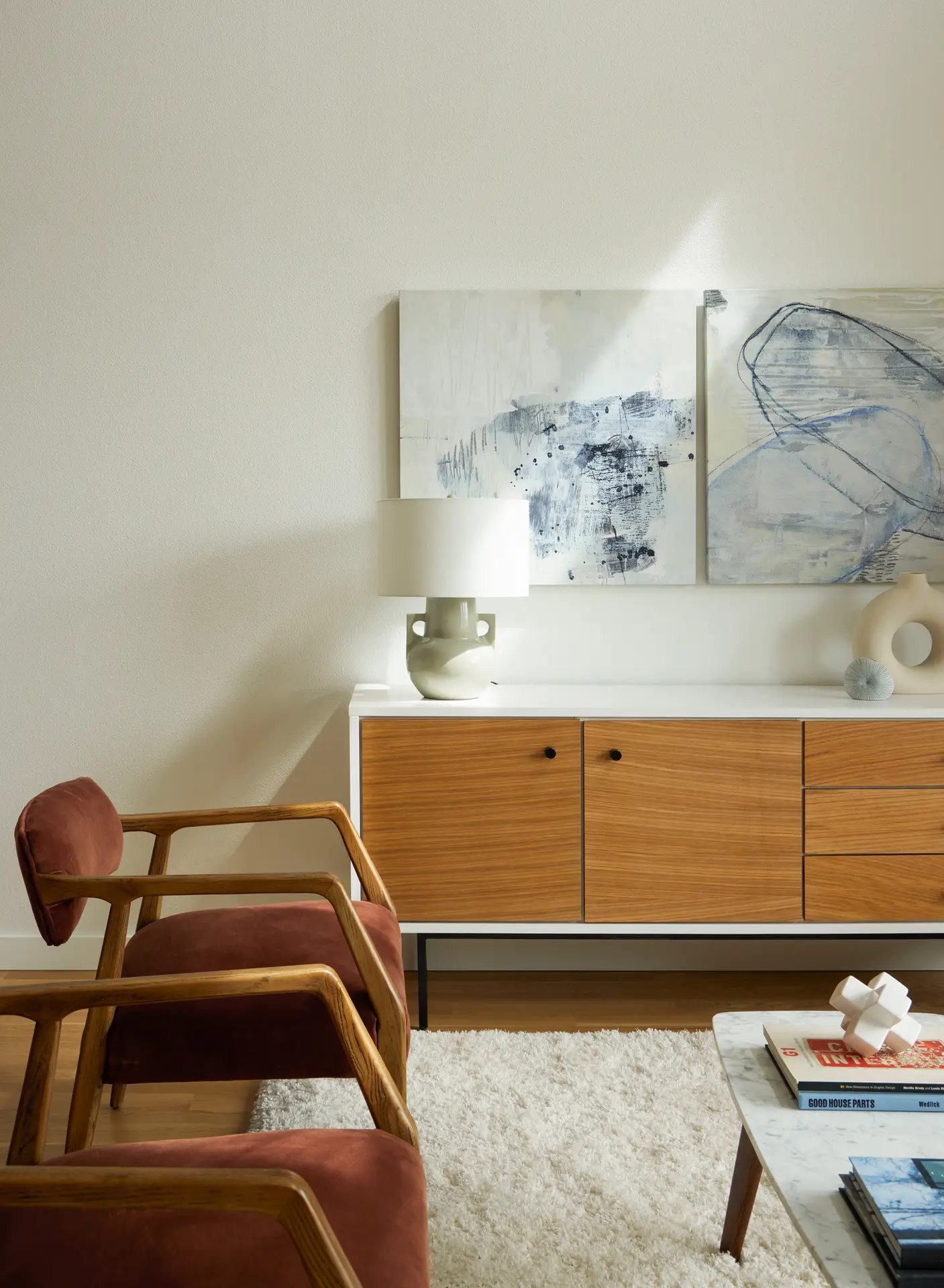 A modern, mid-century-inspired living space with a warm and sophisticated color palette. A long minimalist sideboard features a white frame and natural wood doors and drawers with small black pulls. It sits on slim black metal legs, contributing to the airy feel of the room.
