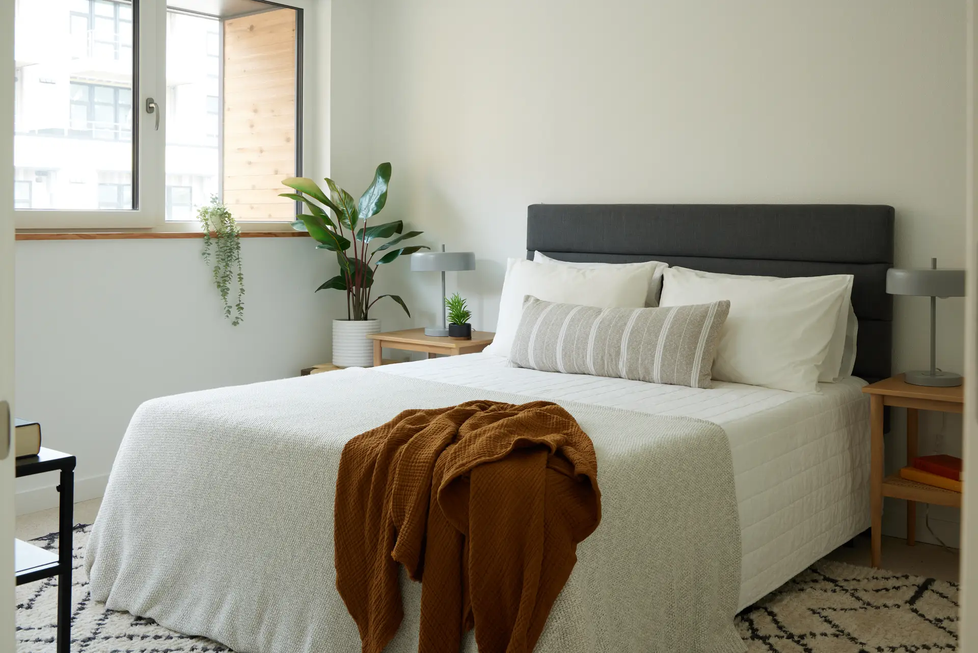 A bedroom at Trillium Townhomes with a bed styled with a white quilted bedspread, white pillows, and a long beige-and-white striped lumbar pillow. A textured burnt orange throw blanket is draped across the foot, adding a pop of warm color to the neutral palette. The room is decorated with several indoor plants, including a large potted bird of paradise and a trailing succulent on the windowsill. A plush, high-pile cream shag rug with a black diamond pattern adds soft texture and warmth to the light-wood floors.