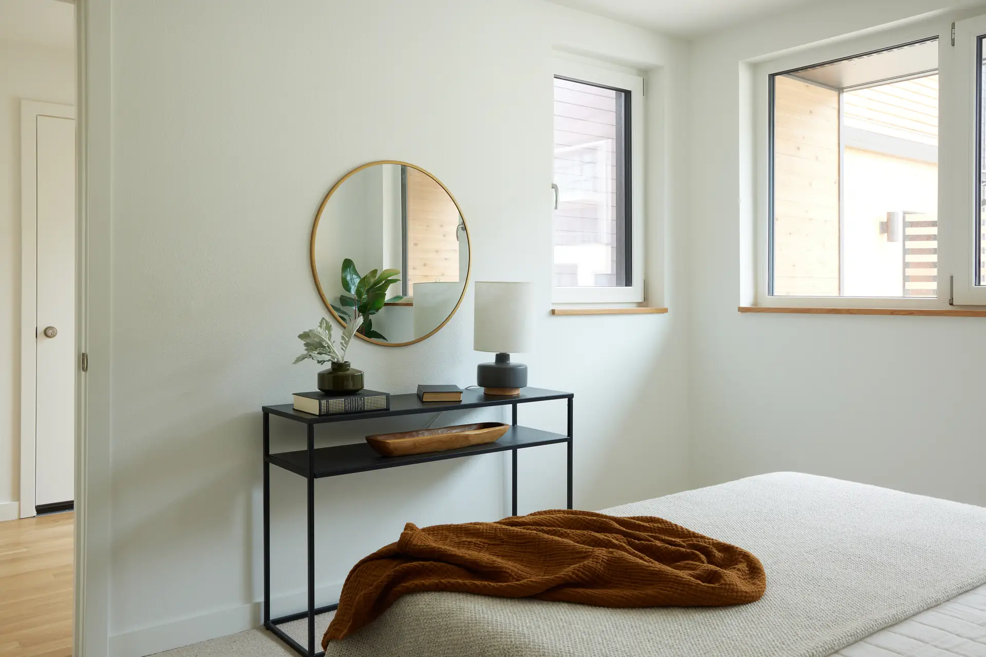 A view of the bedroom with the tilt turn windows and views to the courtyard in the background. A sleek, black metal two-tier console table sits against the wall, serving as a functional display area for a ceramic lamp, books, and a long wooden bowl.
