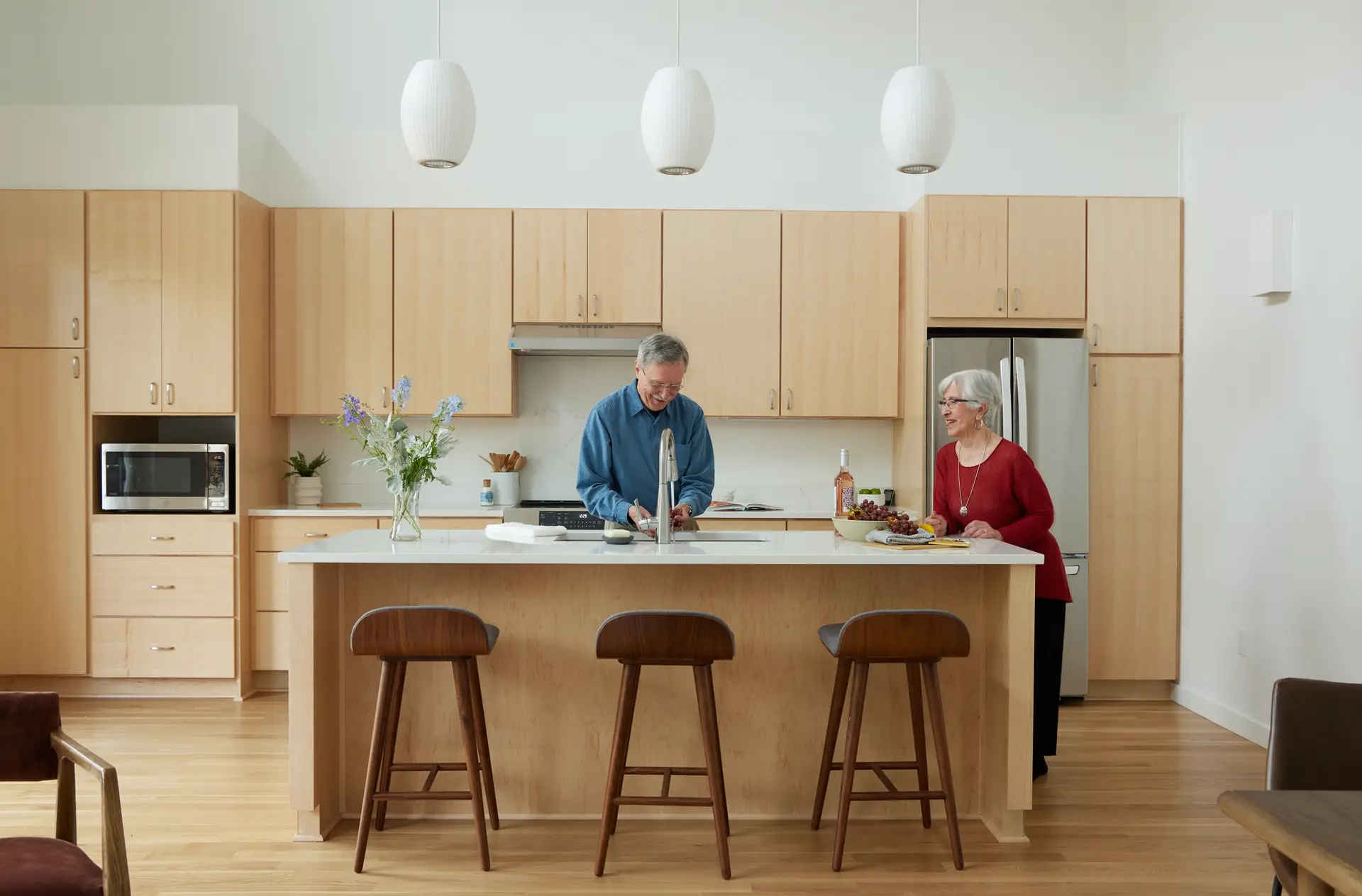 A open-concept kitchen and dining area of a residence at Trillium Townhomes. The space features a clean, contemporary design with light-toned wood cabinetry, likely maple or white oak, and white quartz or stone countertops. Kitchen Island: A central island serves as a focal point, equipped with an undermount sink and three mid-century modern wooden bar stools. Lighting: Three large, white globe pendant lights hang over the island, complementing the airy feel of the room's high ceilings.