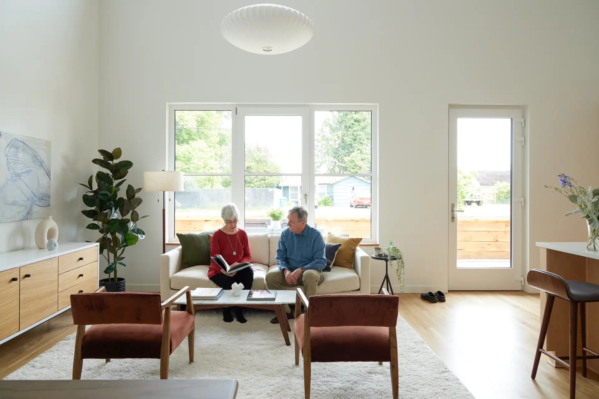Two residents are seated on a low-profile cream sofa, looking at a book together. The area is anchored by a plush, high-pile white area rug and two rust-colored velvet armchairs in the foreground. Behind them is a view to private entry courtyard.
