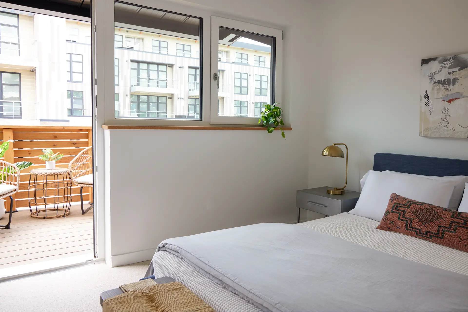 A bedroom, with an open door the second floor balcony outdoor balcony featuring a wood-slat privacy fence, a woven chair, and a small metal bistro table, providing a seamless indoor-outdoor connection.