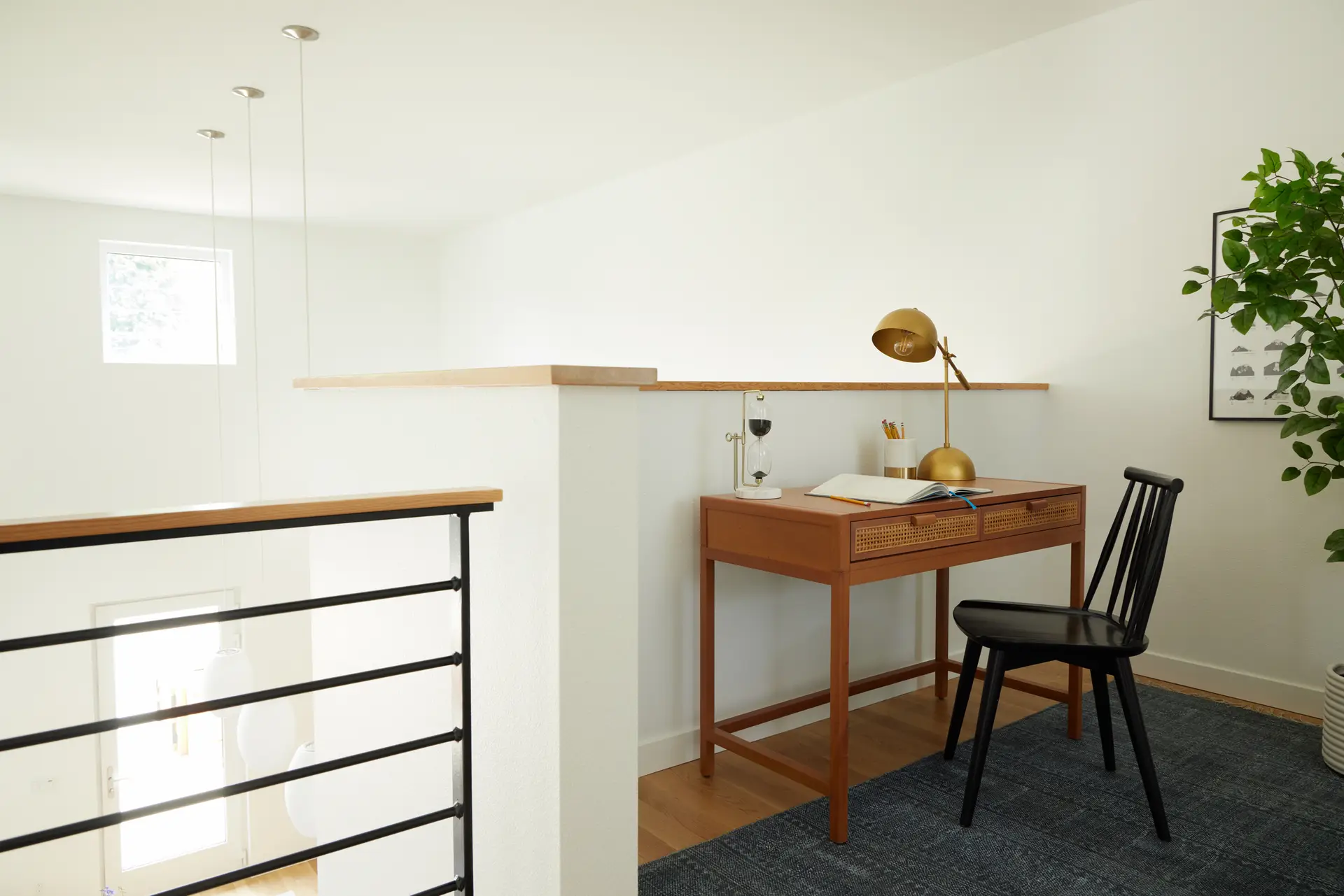 A loft workspace with a slim mid-century modern wooden desk features two drawers with woven cane fronts. It is paired with a black spindle-back chair and a large gold dome-style task lamp. The loft is enclosed by a white pony wall topped with a light-wood ledge, overlooking the home's double-height living area. A black metal horizontal-rail system with a wood handrail lines the staircase landing to the left.