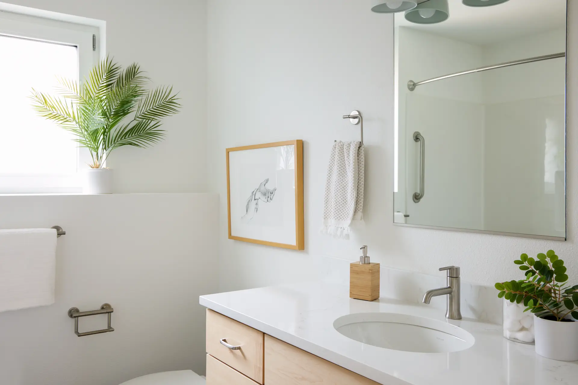A bright, contemporary bathroom from the Trillium Townhomes. The centerpiece is a light-colored white oak vanity with a clean horizontal grain and minimalist silver pulls. It is topped with a thick, crisp white quartz countertop featuring an oval undermount sink and a sleek brushed-nickel faucet. Natural Lighting: A large, high-placed window floods the room with natural light, highlighting the airy atmosphere and white walls.