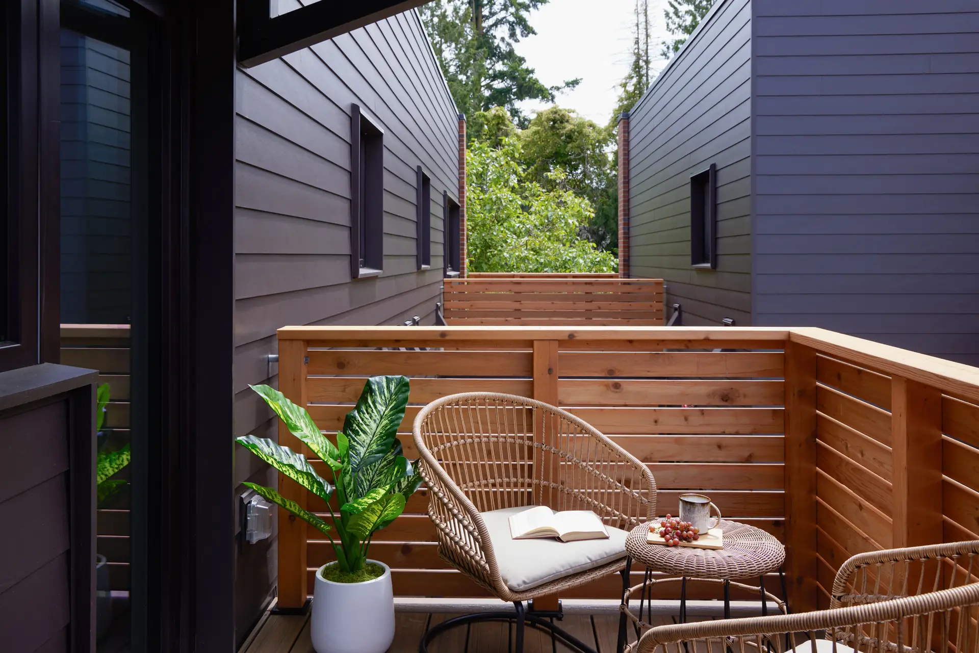 A private outdoor balcony at Trillium Townhomes. The balcony features a warm, clear-stained horizontal wood-slat railing that provides privacy while allowing light and air to pass through. It overlooks a series of similar, modern structures characterized by dark-grey horizontal siding.