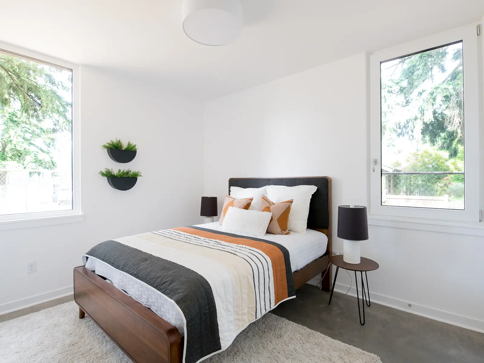 A bright, minimalist bedroom within Going Street Commons. The room features large triple-pane windows that provide abundant natural light while maintaining a superior thermal envelope for energy efficiency and sound insulation. The bed is styled with a modern quilt in grey, orange, and white stripes.
