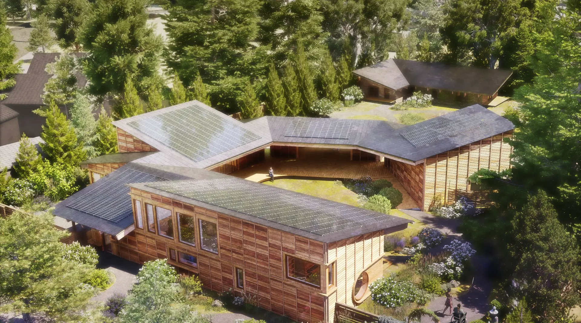 A rendering depicting the proposed expansion of Prismid Sanctuary, a community-driven arts facility and urban retreat in the St. Johns neighborhood of North Portland, Oregon. The facility is designed to be highly sustainable, featuring extensive rooftop solar panels and a super-insulated building envelope. The rendering shows low-profile buildings with natural wood cladding and large windows, designed to harmonize with the surrounding forest and landscape. The buildings are arranged around a central green space, which is intended to serve as a hub for community gatherings, storytelling, and outdoor performances.