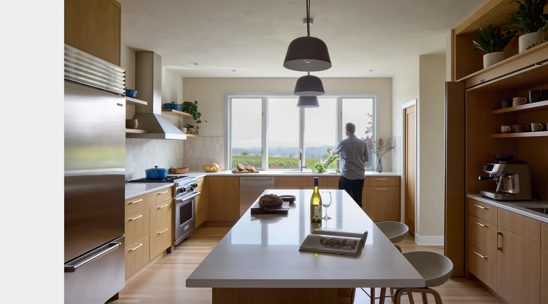 A modern, minimalist kitchen designed & build by Green Hammer. The kitchen features warm wood tones and clean white surfaces. The layout centers around a large island, emphasizing functionality and a connection to the outdoors. The cabinets are made of a light, warm-toned oak wood paired with sleek, elongated gold hardware. The focal point is the kitchen sink infront of an expansive window lookint out to the vineyard beyond. To the right, a dedicated built-in coffee nook features an espresso machine, open shelving for mugs, and potted greenery, neatly integrated into the cabinetry.