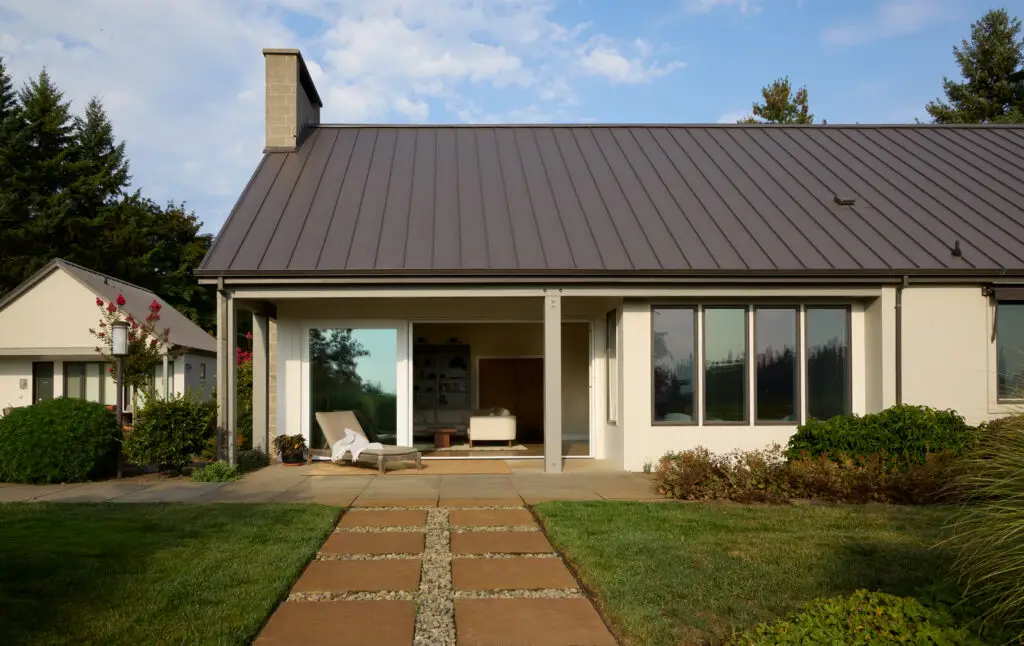 The back of the home from the terrace with the sliding door open to see into the living room. The home has a dark standing seam metal roof, succo cladding and exposed wood posts and beams. The pool house is visable to the left.