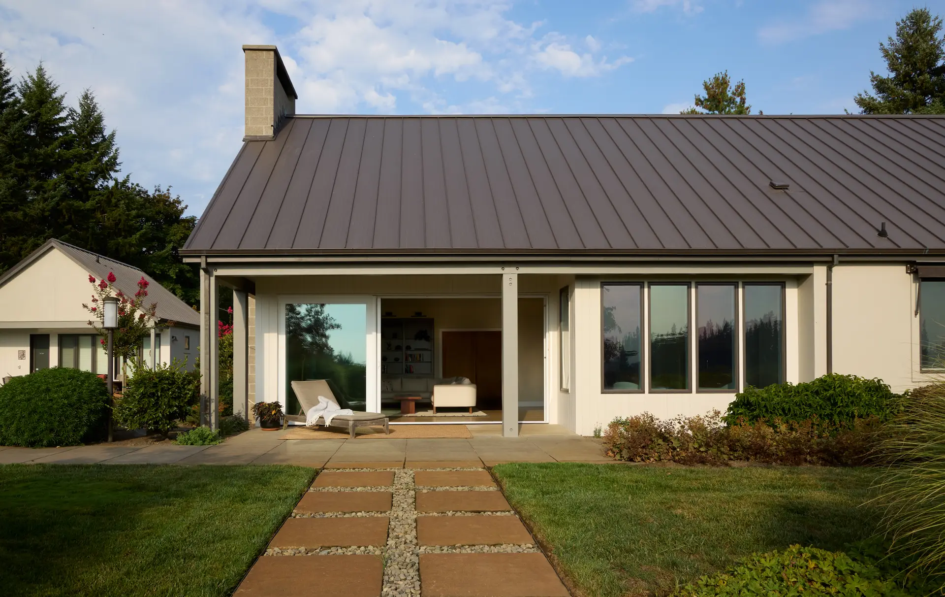 The back of the home from the terrace with the sliding door open to see into the living room. The home has a dark standing seam metal roof, succo cladding and exposed wood posts and beams. The pool house is visable to the left.