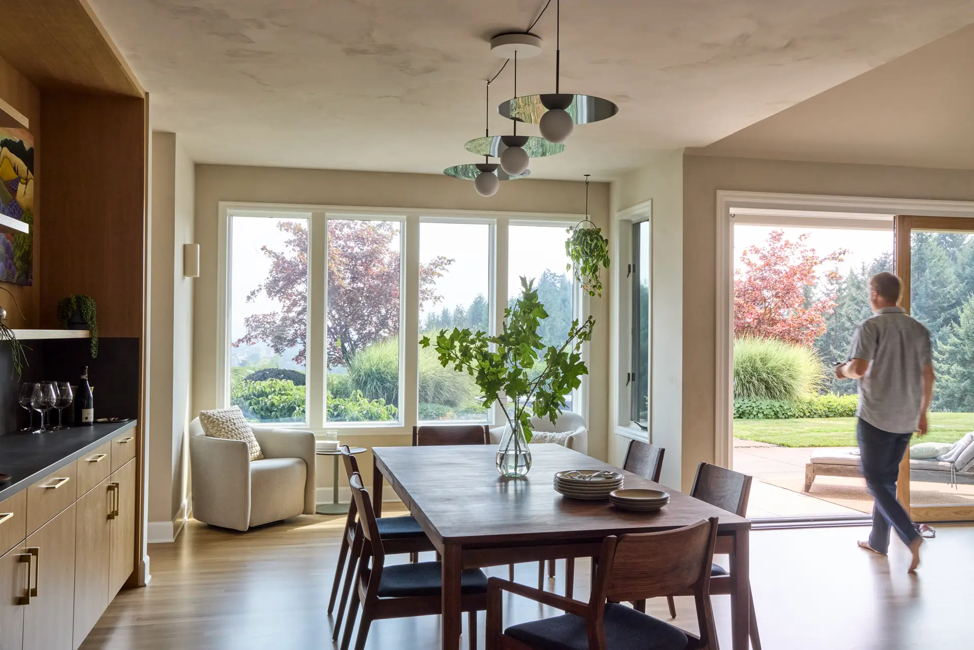 The dining room looking toward the vineyard and the open sliding door on the left. In the center is a custom table by The Jointery, in Portland Oregon. A pendant fixture with three forsted round lamps with mirrored reflectors hangs above the table. On the left is the bar.