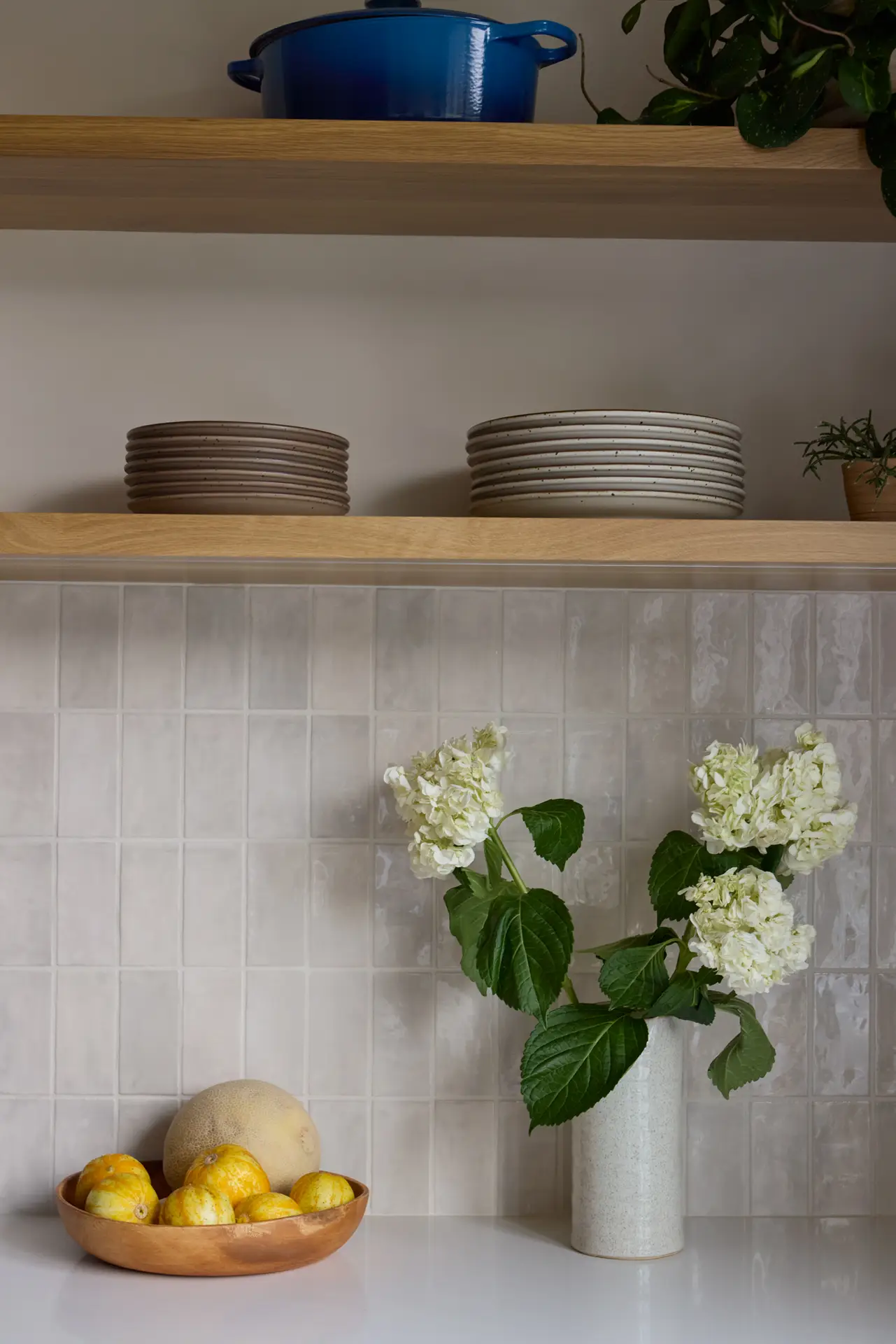 Close-up view of the kitchen backspalsh tile. The tile is installed in a vertical stack-bond with a iridescent glaze. White oak floating shelves above hold dishware.