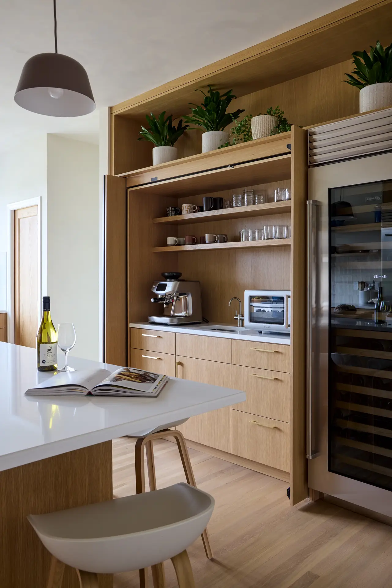 A "hidden" coffee and beverage bar behind light oak cabinetry doors that slide back into "pockets" on either side, allowing the station to be fully opened during use and completely hidden when not in use. To the right is a professional-grade Sub-Zero wine refridgerator with a glass front and stainless steel trim.