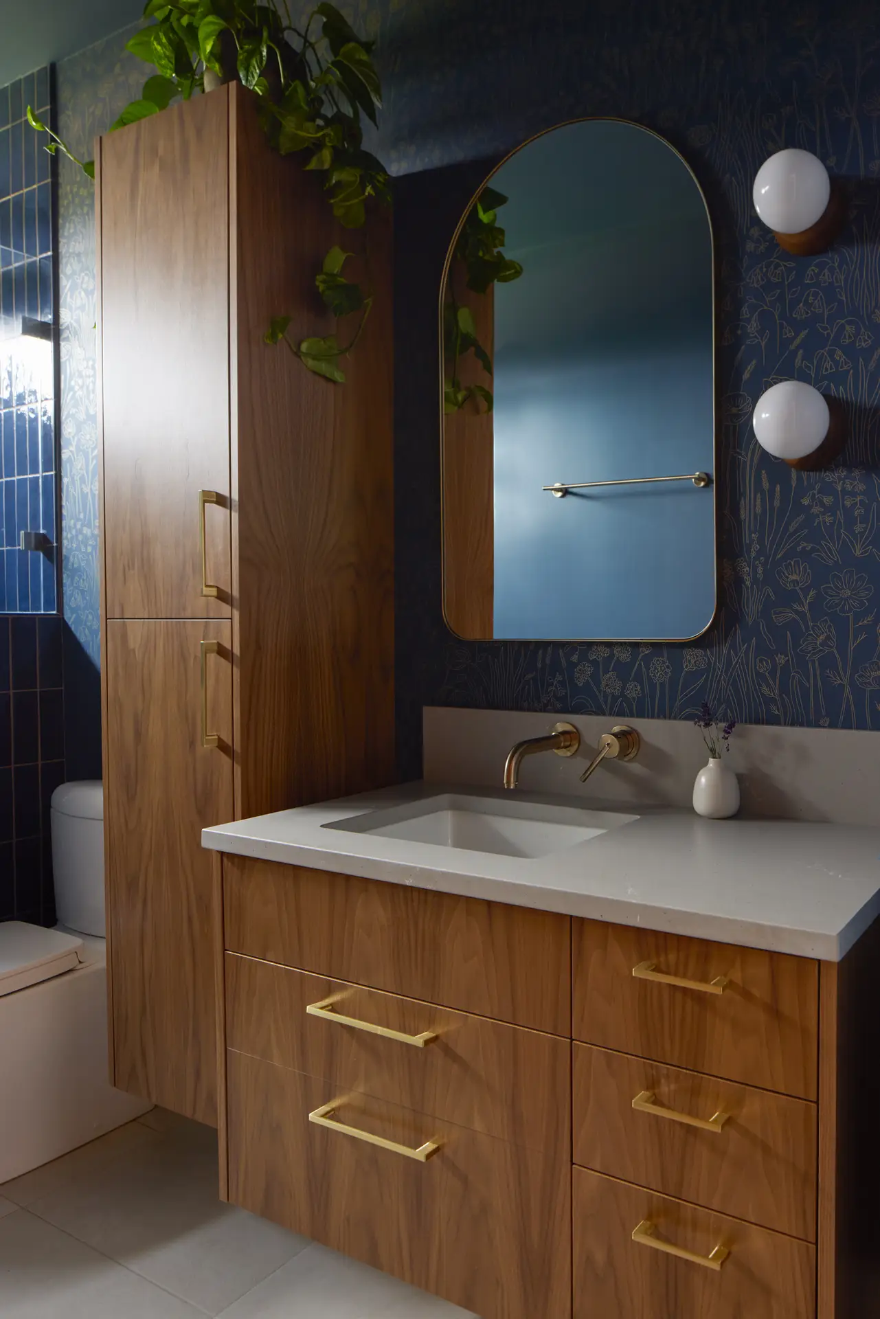 The guest bathroom wall hung vanity with a matching tall linen cabinet. The wall features a dark navy floral wallpaper with gold biophlic patterns. Above the sink is a large arched mirror with a thin gold frame.