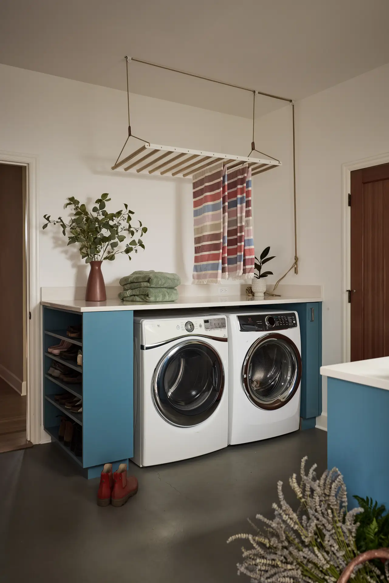 The laundry room with a side by side washer and drywer with a countertop above and open shoe storage to the left. A ceiling mounted drying rack lowers and raises via a pully system. The floor is painted a dark grey.