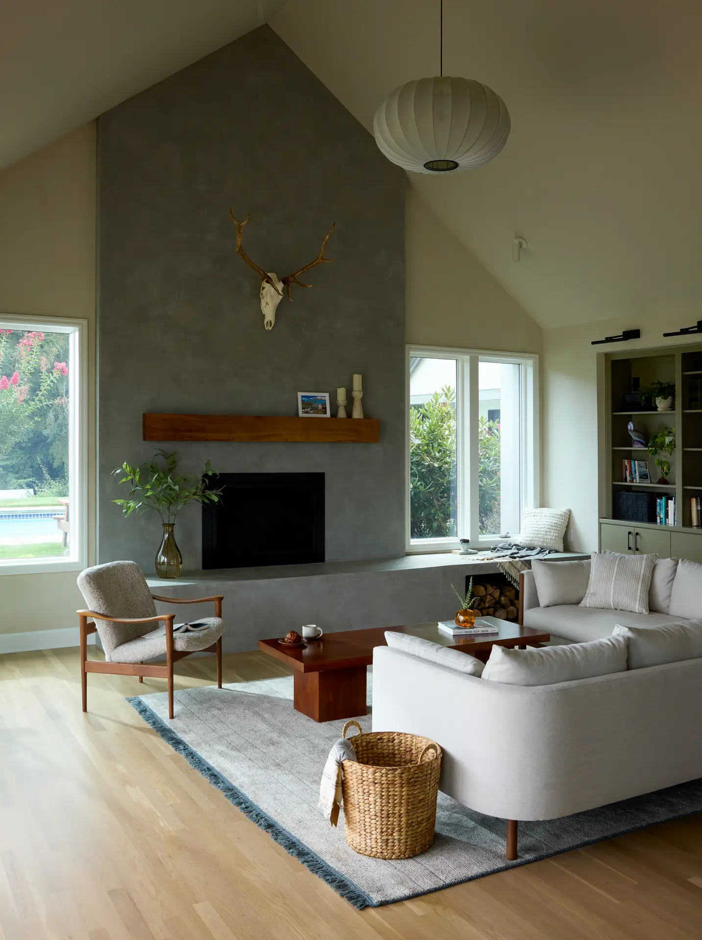 A spacious living room with a microcement clad fireplace as the centerpiece. Above the fireplace is a thick, floating reclaimed wood mantel. There are two windows on each side fo the fireplace with views to the pool. On the left is a built-in bookcase. A plush, off-white sectional sofa from Design Within Reach anchors the living area, complemented by a mid-century modern wooden armchair with light-colored upholstery.