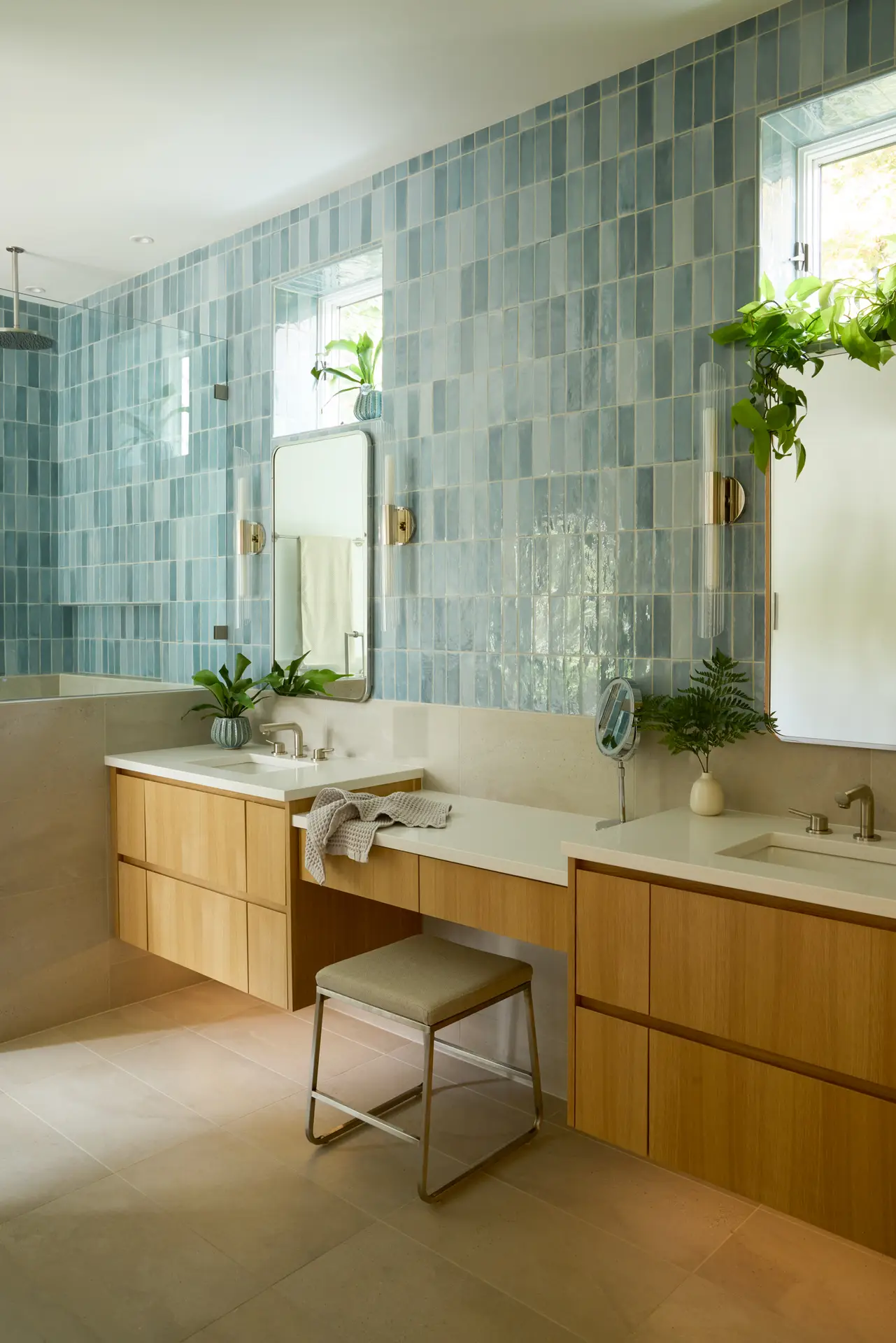 Primary suite emphasizing a bright, airy aesthetic with natural sunlight and a blend of organic and modern materials. The focal point is the floating vanity and makeup station, which is crafted from white oak by Big Branch Woodworking. Behind the wall and shower are clad in ceramic tiles from Pratt + Larson. Above each sink is a mirrored medicine cabinet flanked by wall sconces.