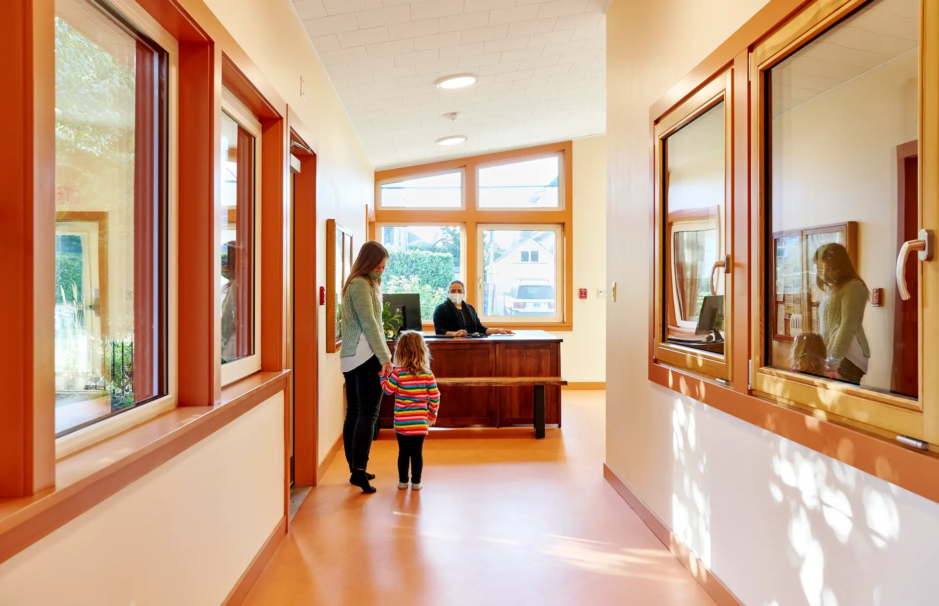 New Day School, showing a mother and child approaching the reception desk. Beyond the reception is a lovely window which lets plenty of light into the space. Softly textured cork floors combined with wood windows and doors create a warm and welcoming atmosphere.