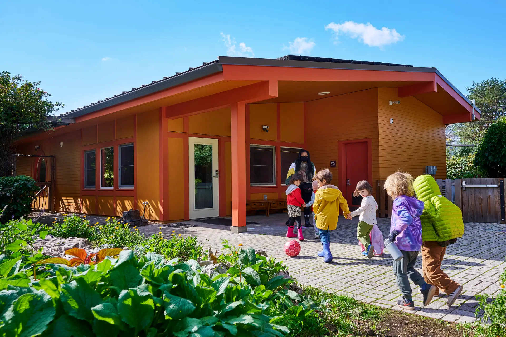 Children entering the New Day School which has bright orange siding and a coral trim. The building is constructed in a craftsman style which reflects the character of the neighborhood.
